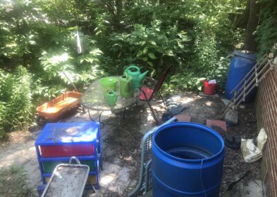 Wildergarden patio with two blue rain barrels, a red bucket a red wagon, a plastic set of drawers for catching rain, and a table with three bright green watering cans, and a red chair, Surrounding the patio is a forest of herbaceous plants, shrubs, and trees.