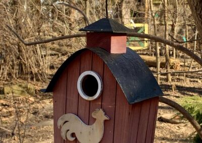 Wooden birdhouse built to resemble a barn. There is the outline of a rooster wind vane on the front, below the hole birds can use to enter. The birdhouse is hanging from a tree in the fall, so there are no leaves on the trees in the background.