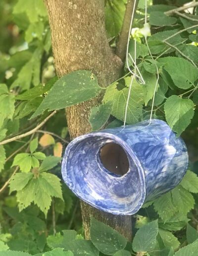 Ceramic birdhouse shaped like a tube that is closed at one end and mostly closed at the other, but with a round hole for birds to get in. The glaze is a patchy dark blue. The birdhouse is hanging in from a tree with smooth bark and is surrounded by dark green leaves.