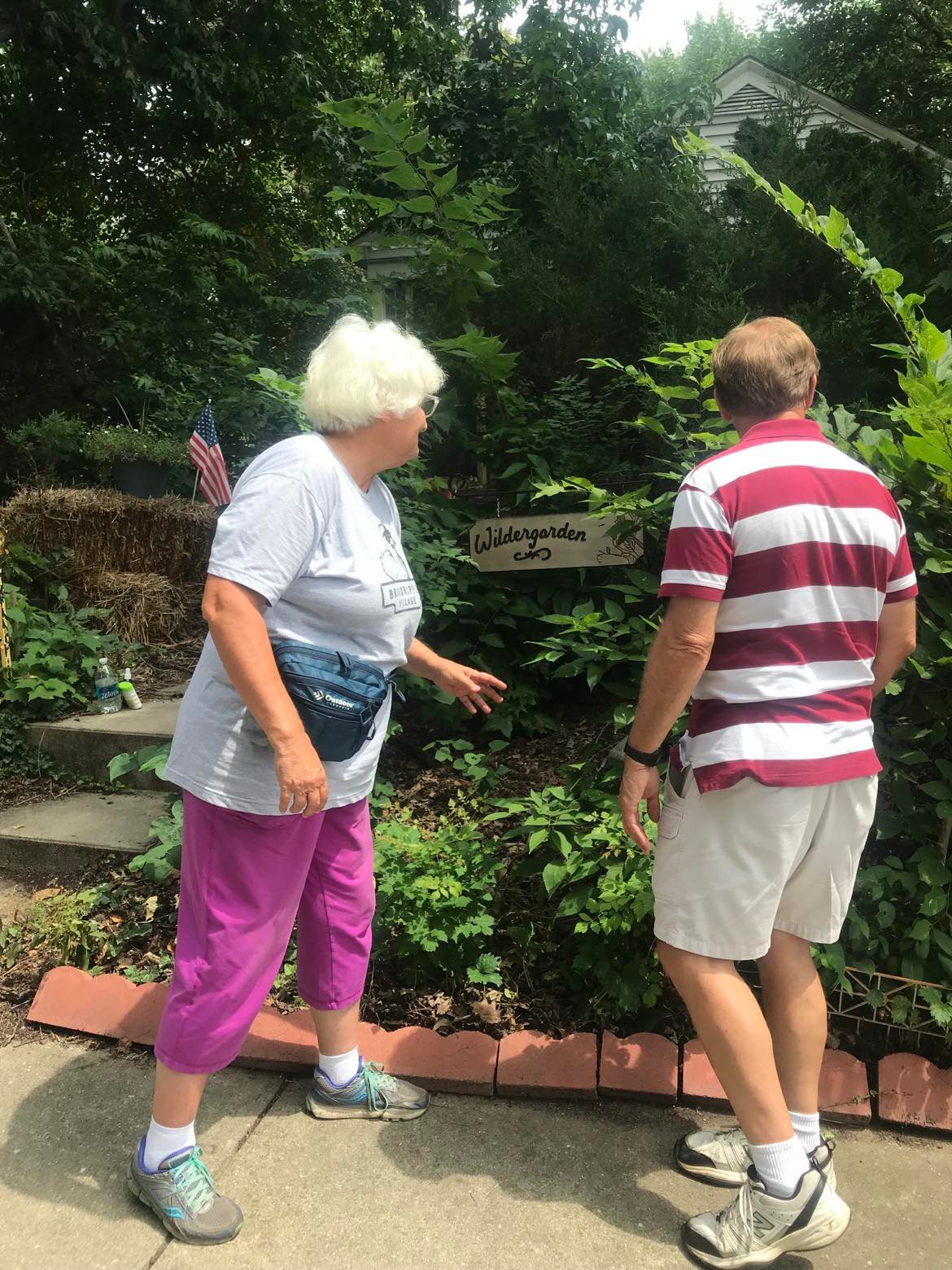 Building Community! Julia, a vigorous, gray-haired woman wearing a lavender shirt, pink pants, and a blue bag speaks to her friend Rex, a man in a red and white striped shirt and white shorts, while standing in front of the Wildergarden sign. There are many plants of all sizes, and they almost completely obscure the white garage in the background.