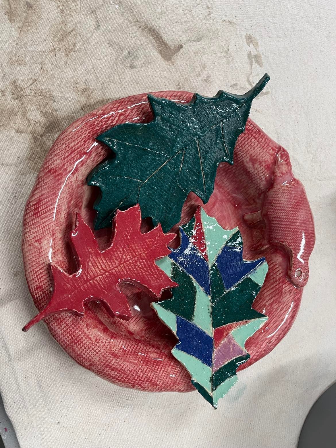 Art in the Garden! Handmade reddish-brown ceramic bowl with three brightly colored clay leaves across the top. Leaves are solid green, solid red, and an artistic patchwork of red, dark blue, dark green, and light blue. It catches rainwater for wildlife to drink.