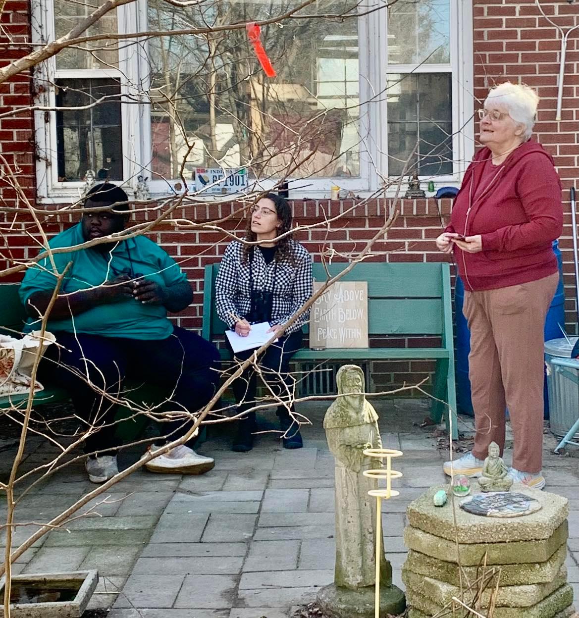 Two people sit on green benches on the Wildergarden porch, while Julia stands. All three are gazing in the same direction as they attempt to identify birds visually and using the Merlin phone app.