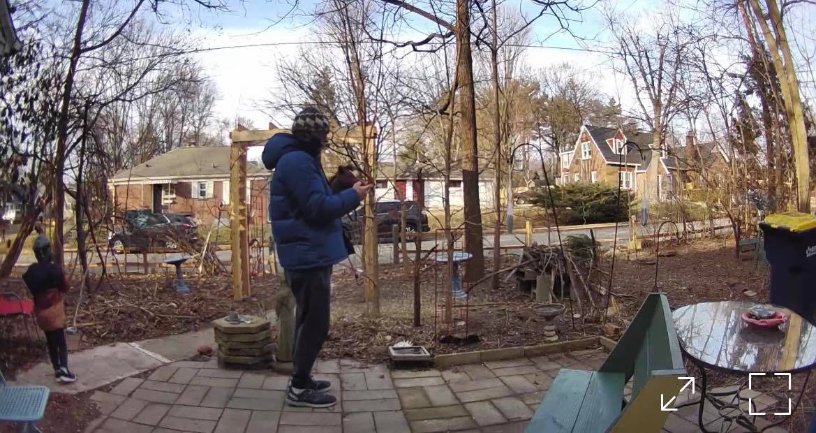 A man in a bright blue winter coat stands near his son on the Wildergarden patio in winter when all the trees have lost their leaves and all the plants have died back for the winter, so you have a clear view of the street in the background.