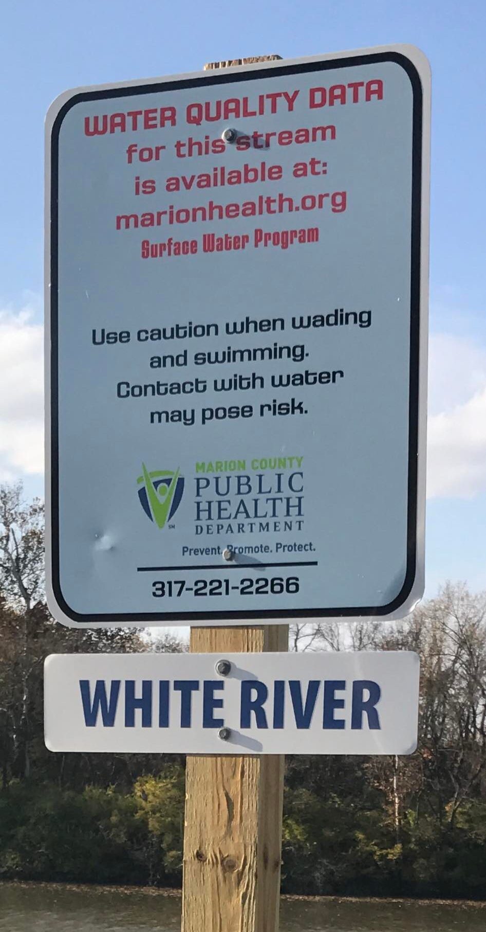 Sign posted in front of the White River that reads: Water quality data for this stream is available at marionhealth.org Surface Water Program Use caution when wading and swimming. Contact with water may pose risk. Marion County Public Health Department Prevent. Promote/ Protect. 317-221-2266